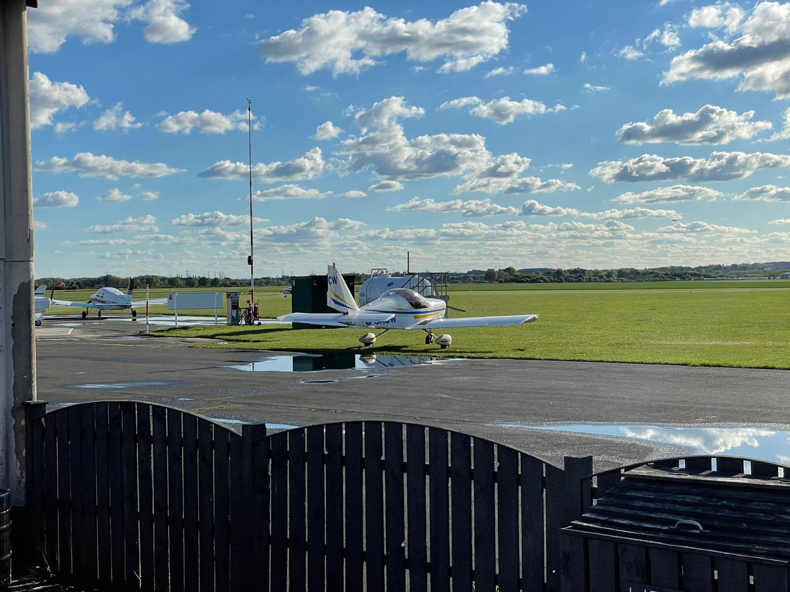 Two light aircraft parked on an airfield apron under a wide blue sky with scattered cloud.